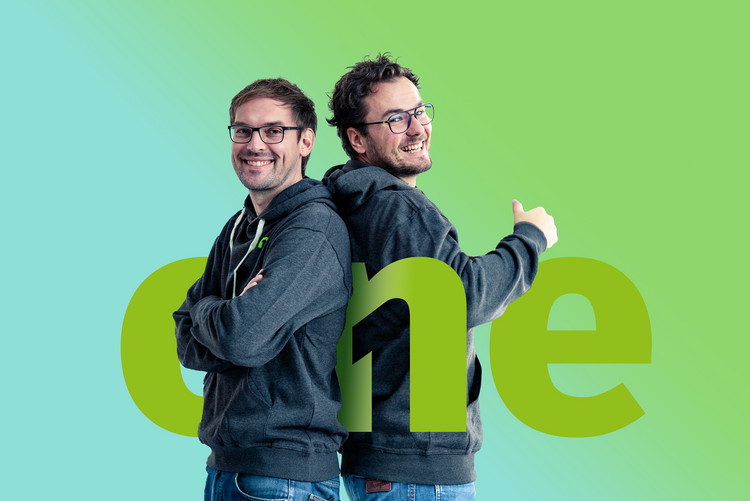 Zwei Männer stehen Rücken an Rücken vor grünem Hintergrund und lachen in die Kamera, zwischen ihnen steht das große Logo von destination.one