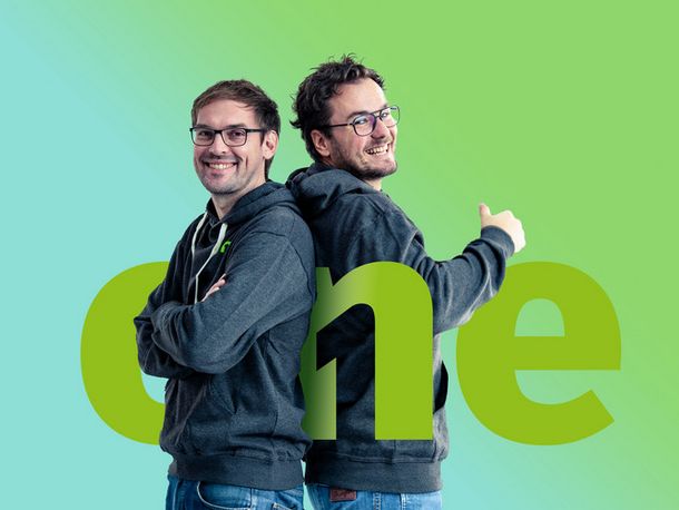 Zwei Männer stehen Rücken an Rücken vor grünem Hintergrund und lachen in die Kamera, zwischen ihnen steht das große Logo von destination.one