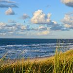 Blick über eine Düne und Strand auf das offene Meer mit blauem, leicht bewölktem Himmel
