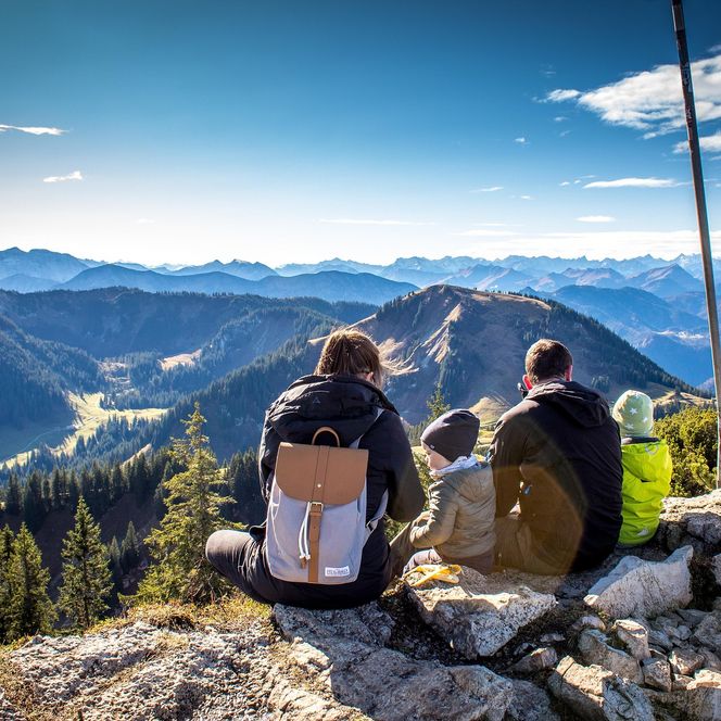 Familie mit zwei Kindern sitzt auf einem Berggipfel und schaut in den Horizont mit weiteren Bergen