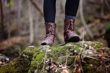 Person in braunen Lederstiefeln steht im Wald auf einem Stein mit Moos 