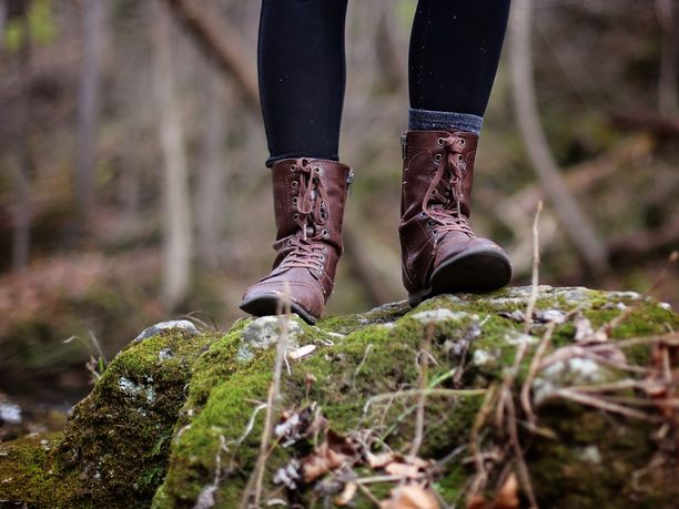 Person mit braunen Lederstiefeln steht auf einem moosigen Felsen im Wald.