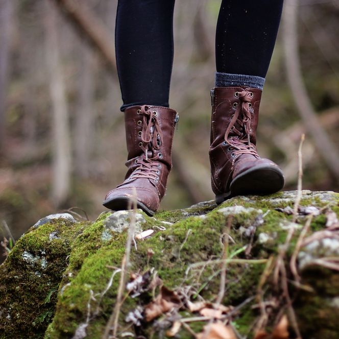 Person mit braunen Lederstiefeln steht auf einem moosigen Felsen im Wald.